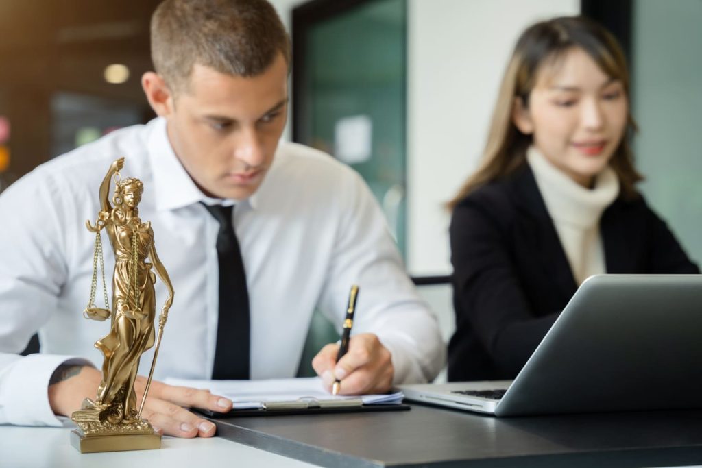 Un abogado hombre y una abogada mujer trabajando en documentos con una estatua de la Dama de la Justicia en primer plano.